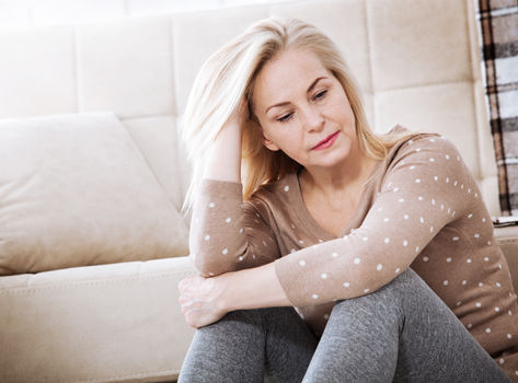 Thoughtful-looking woman sitting on the floor near sofa, embracing her knees