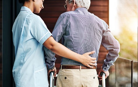 A nurse puts her arm around an elderly male patient as he walks with a walker.